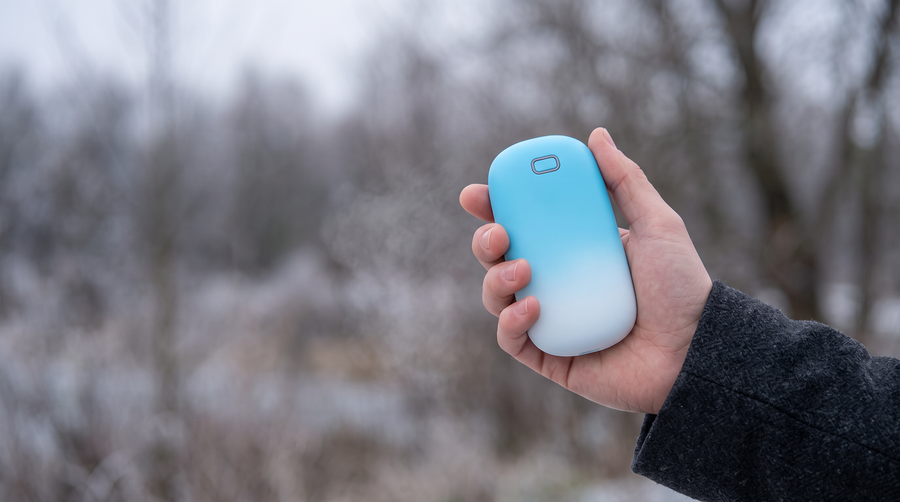 The blue hand warmer held in a bare hand outdoors on a frozen winter day, filling the palm, with frosted branches out of focus behind.